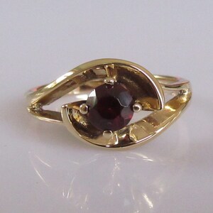 May include: A gold ring with a dark red gemstone set in a swirling design.