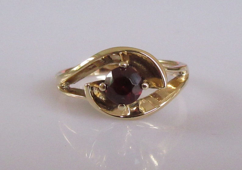 May include: A gold ring with a dark red gemstone set in a swirling design.