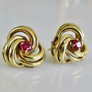 May include: A pair of gold earrings with a red ruby stone in the center of each earring. The earrings are in the shape of a knot.