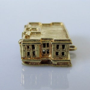 Puede incluir: Un amuleto de color dorado en forma de edificio con techo plano y ventanas. El amuleto tiene un bucle en el lateral para sujetarlo a una pulsera o collar.