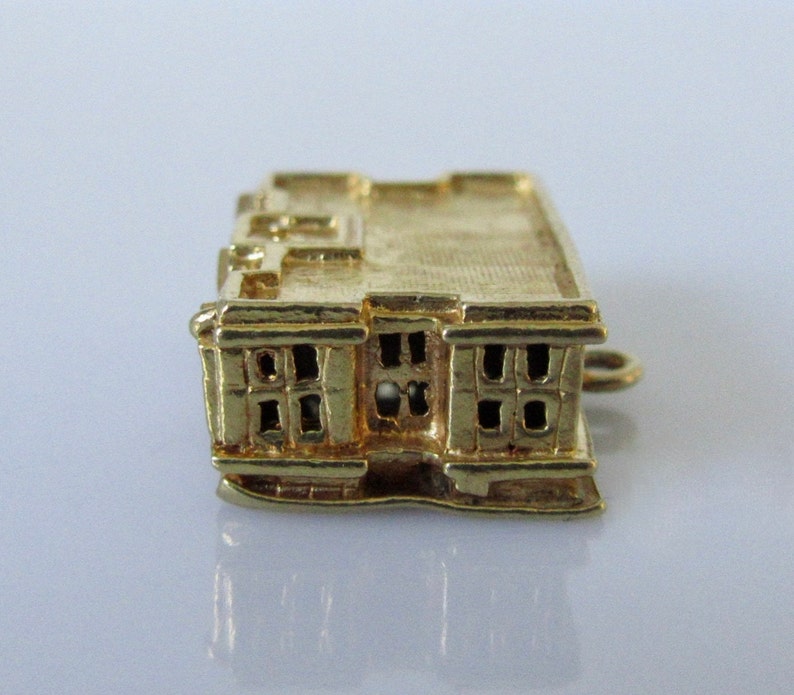 Puede incluir: Un amuleto de color dorado en forma de edificio con techo plano y ventanas. El amuleto tiene un bucle en el lateral para sujetarlo a una pulsera o collar.