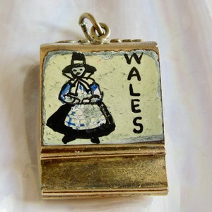 Pu&ograve; includere: Ciondolo in oro con un'immagine dipinta di una donna in costume tradizionale gallese. Il ciondolo ha una cornice dorata e la parola "WALES" &egrave; scritta verticalmente sul lato destro.