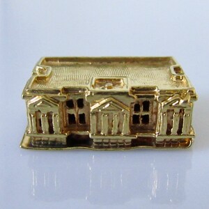 Puede incluir: Un amuleto de edificio en miniatura de color dorado con una fachada detallada y un techo plano.