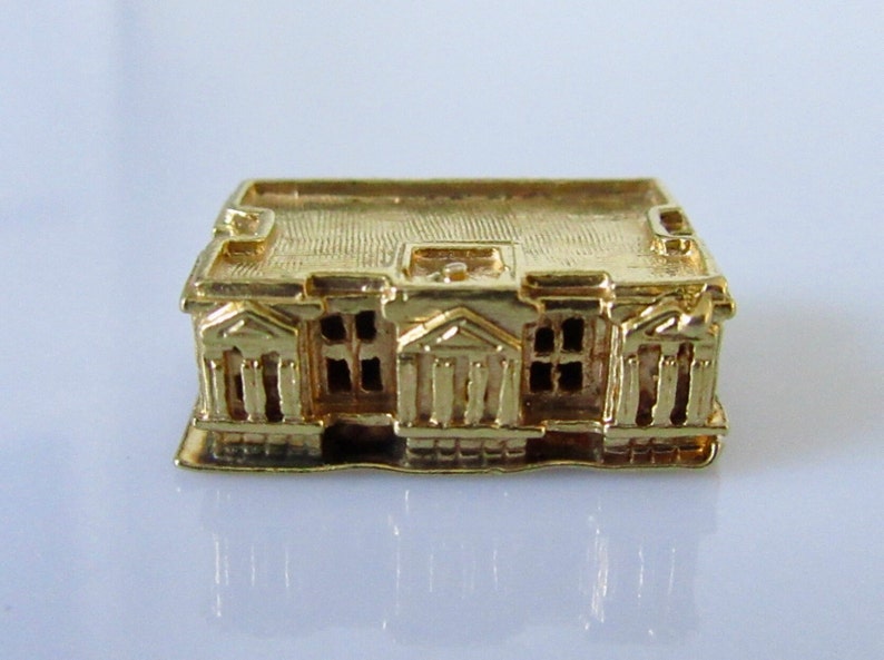 Puede incluir: Un amuleto de edificio en miniatura de color dorado con una fachada detallada y un techo plano.