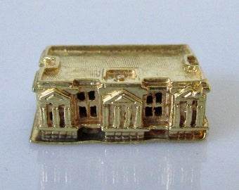 Può includere: Un ciondolo a forma di edificio in miniatura color oro con una facciata dettagliata e un tetto piatto. L'edificio ha un design simmetrico con più finestre e colonne.
