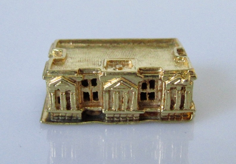 Puede incluir: Un amuleto de edificio en miniatura de color dorado con una fachada detallada y un techo plano. El edificio tiene un dise&ntilde;o sim&eacute;trico con m&uacute;ltiples ventanas y columnas.