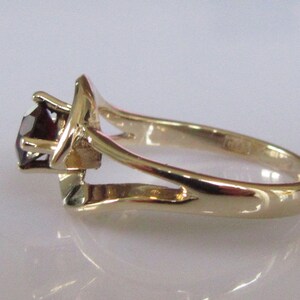 May include: A gold ring with a single, dark red gemstone set in a prong setting. The ring has a delicate, open-work design.