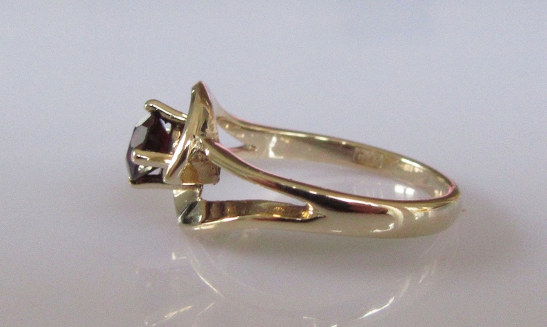 May include: A gold ring with a single, dark red gemstone set in a prong setting. The ring has a delicate, open-work design.
