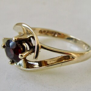 May include: A gold ring with a red gemstone set in a prong setting. The ring has a unique, modern design with a curved band.