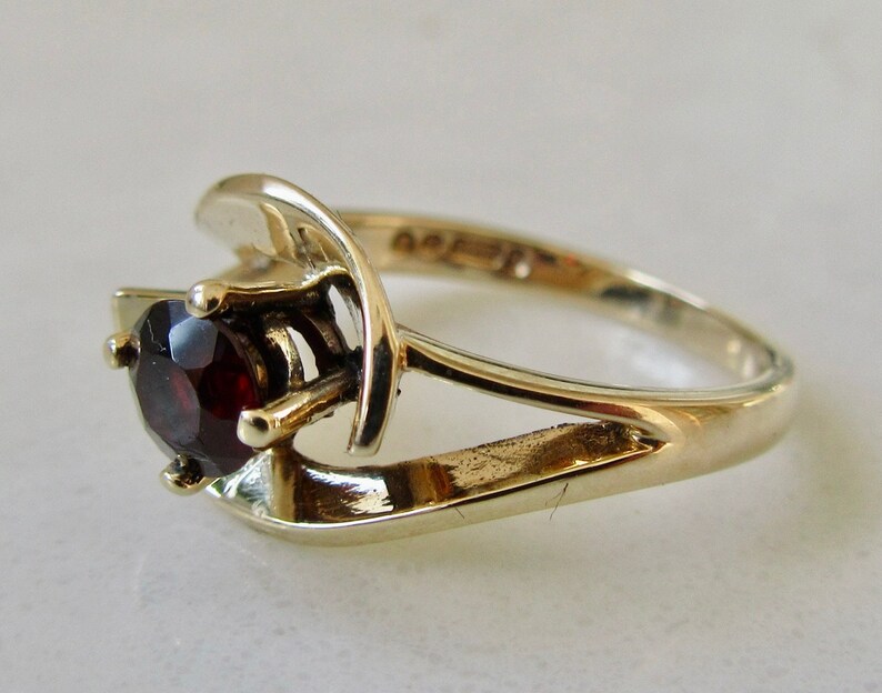 May include: A gold ring with a red gemstone set in a prong setting. The ring has a unique, modern design with a curved band.