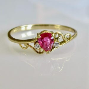 Może przedstawiać: Złoty pierścionek z różowym, gruszkowatym kamieniem szlachetnym i dwoma mniejszymi białymi kamieniami osadzonymi w delikatnym, wijącym się wzorze.