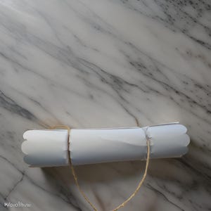 May include: A white paper tube with scalloped edges is tied with twine. The tube is lying on a white marble surface.