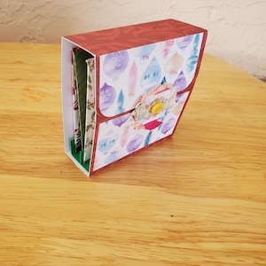 May include: A handmade paper craft box with a red patterned top and a flap decorated with Christmas ornament designs. The box features a floral embellishment and a yellow button closure. The box is on a wooden surface.