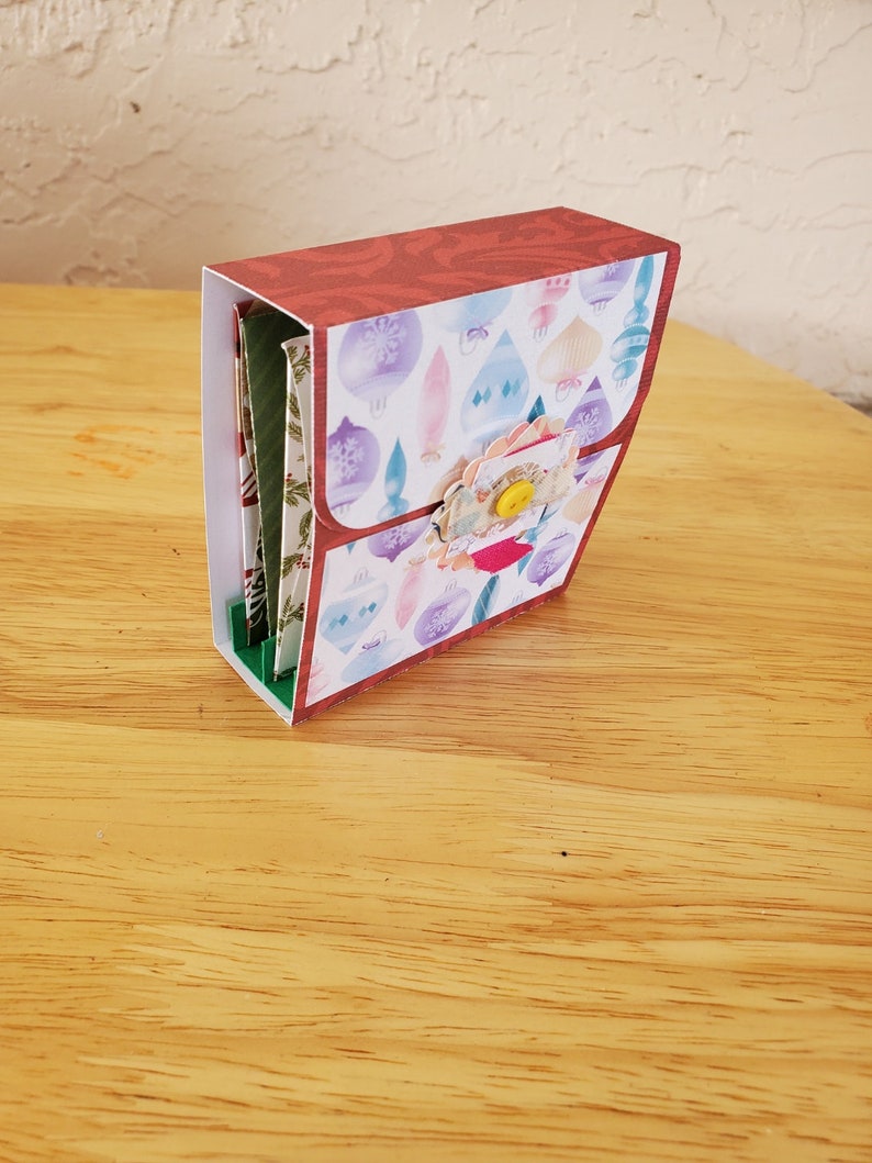 May include: A handmade paper craft box with a red patterned top and a flap decorated with Christmas ornament designs. The box features a floral embellishment and a yellow button closure. The box is on a wooden surface.