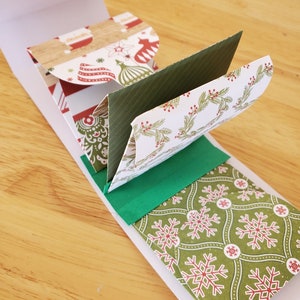 May include: A festive Christmas card with a white outer sleeve and multiple folded interior panels. The panels display seasonal patterns, such as snowflakes, baubles, and Santa Claus, in red, green, and white. The card is open, showcasing the layered design.
