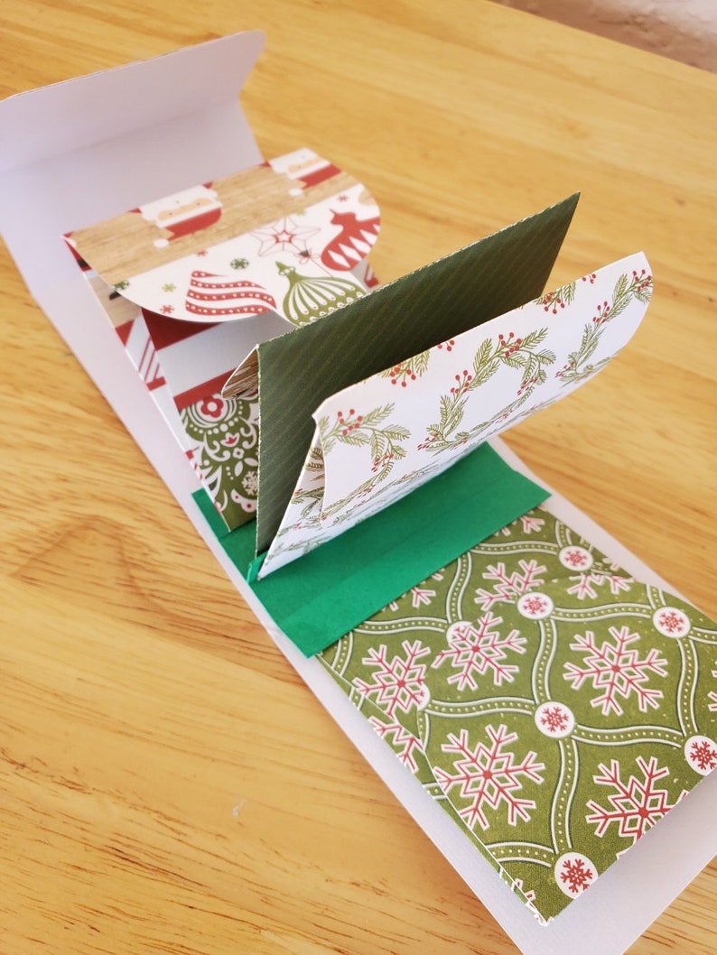 May include: A festive Christmas card with a white outer sleeve and multiple folded interior panels. The panels display seasonal patterns, such as snowflakes, baubles, and Santa Claus, in red, green, and white. The card is open, showcasing the layered design.
