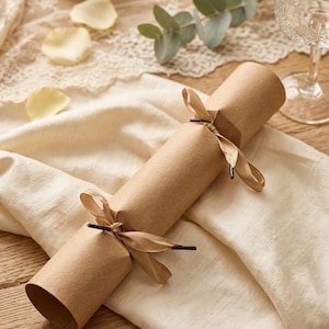 May include: A cylindrical party cracker wrapped in plain brown kraft paper, secured at both ends with light tan satin ribbons tied into bows. The cracker features a simple, unadorned design with a hollow opening at the visible end, showcasing the paper texture.