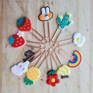 Puede incluir: Una colección de clips decorativos con dijes coloridos de estilo caricatura. Los dijes incluyen una fresa, un conejito, un cactus, una flor, un arcoíris, una piña, una cereza y un plátano. Los clips están dispuestos en un patrón circular sobre una superficie de madera.