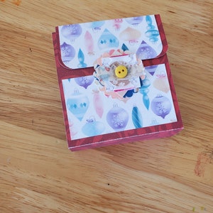 May include: A small, rectangular gift box with a flap closure. The box features a festive design with colorful Christmas ornaments on a white background. A decorative embellishment with a yellow button adorns the front, adding a touch of charm.