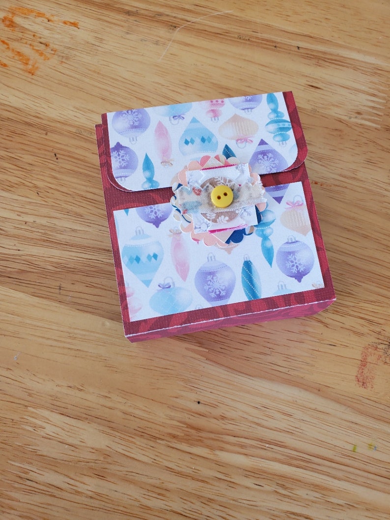 May include: A small, rectangular gift box with a flap closure. The box features a festive design with colorful Christmas ornaments on a white background. A decorative embellishment with a yellow button adorns the front, adding a touch of charm.
