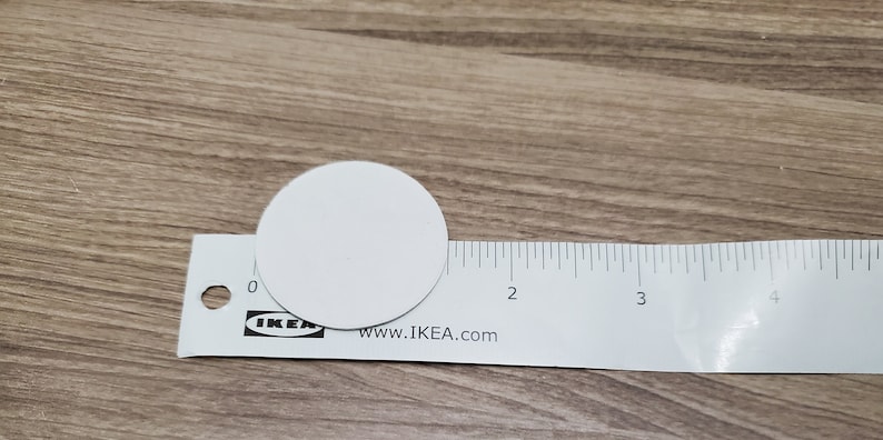 Pode incluir: Um c&iacute;rculo branco com aproximadamente 3,8 cm de di&acirc;metro, colocado em cima de uma r&eacute;gua branca com marcas pretas. A r&eacute;gua &eacute; etiquetada com "IKEA" e "www.IKEA.com".