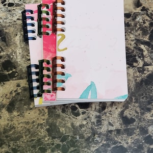 May include: Three spiral bound notebooks with colorful watercolor designs. The notebooks are stacked on top of each other and have plastic binding rings.