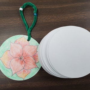 Pode incluir: Um limpador de cachimbo verde com contas brancas e azuis est&aacute; preso a um ornamento redondo de papel com uma flor laranja desenhada &agrave; m&atilde;o sobre um fundo verde. Uma pilha de ornamentos redondos de papel branco est&aacute; ao lado do ornamento.