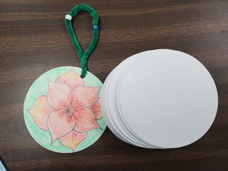 Pode incluir: Um limpador de cachimbo verde com contas brancas e azuis est&aacute; preso a um ornamento redondo de papel com uma flor laranja desenhada &agrave; m&atilde;o sobre um fundo verde. Uma pilha de ornamentos redondos de papel branco est&aacute; ao lado do ornamento.