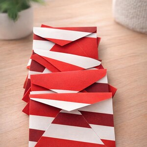 May include: A stack of small, square envelopes with a red and white striped pattern. The envelopes are folded and stacked on a light wooden surface. A potted plant and a white fabric bag are in the background.
