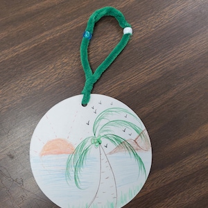 Pode incluir: Um ornamento redondo de papel branco com um cabide de limpador de cachimbo verde. O ornamento apresenta uma ilustra&ccedil;&atilde;o desenhada &agrave; m&atilde;o de uma palmeira numa praia com um p&ocirc;r do sol e p&aacute;ssaros a voar no c&eacute;u.