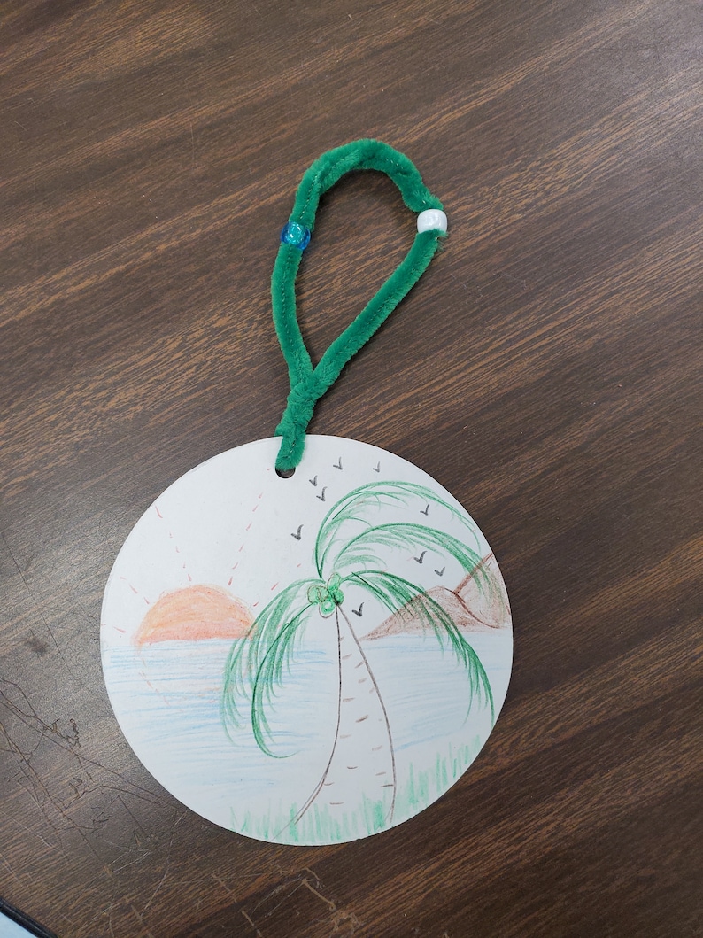 Pode incluir: Um ornamento redondo de papel branco com um cabide de limpador de cachimbo verde. O ornamento apresenta uma ilustra&ccedil;&atilde;o desenhada &agrave; m&atilde;o de uma palmeira numa praia com um p&ocirc;r do sol e p&aacute;ssaros a voar no c&eacute;u.