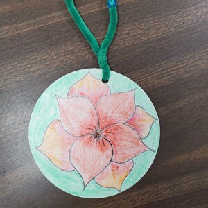 Pode incluir: Um ornamento redondo de papel verde com uma flor vermelha desenhada nele. O ornamento tem um la&ccedil;o de limpador de cachimbo verde preso ao topo.