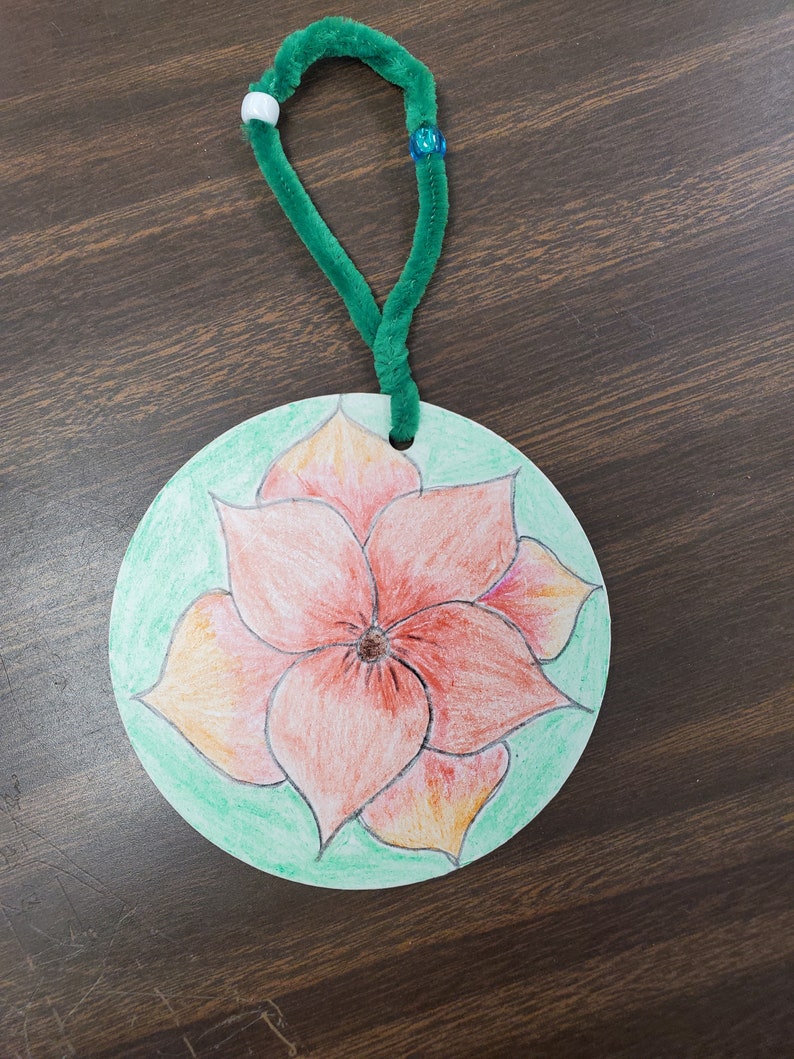 Pode incluir: Um ornamento redondo de papel verde com uma flor vermelha desenhada nele. O ornamento tem um la&ccedil;o de limpador de cachimbo verde preso ao topo.