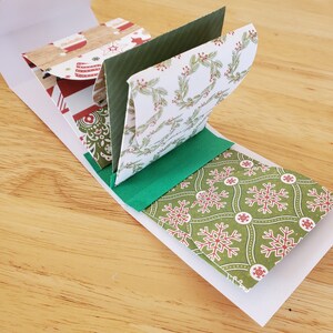 May include: A handmade Christmas card with a white envelope and a pop-up design. The card features festive patterns, including snowflakes, wreaths, and Santa Claus, in red, green, and brown hues. The card is open, showcasing the interior design.