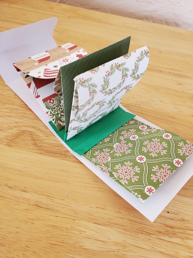 May include: A handmade Christmas card with a white envelope and a pop-up design. The card features festive patterns, including snowflakes, wreaths, and Santa Claus, in red, green, and brown hues. The card is open, showcasing the interior design.
