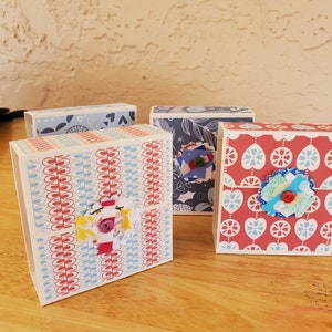 May include: Four decorative paper boxes with patterned designs. The boxes feature red, blue, and white colours. Each box has a unique pattern, including floral and geometric designs. Some boxes are embellished with fabric flowers and buttons.