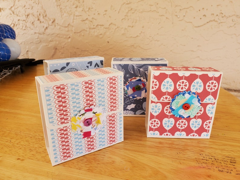 May include: Four decorative paper boxes with patterned designs. The boxes feature red, blue, and white colours. Each box has a unique pattern, including floral and geometric designs. Some boxes are embellished with fabric flowers and buttons.