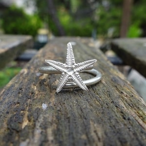 Puede incluir: Un anillo de plata con un diseño de estrella de mar. El anillo está sobre una superficie de madera.