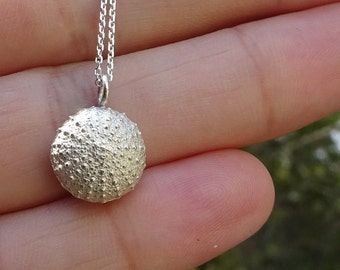 Può includere: Una collana d'argento con un ciondolo a forma di riccio di mare dettagliato. Il ciondolo è testurizzato e ha una forma rotonda.