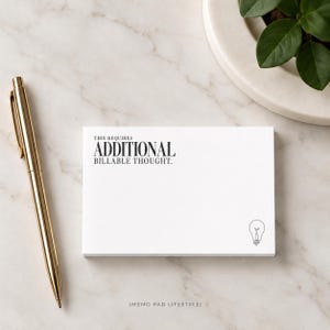 May include: A white rectangular memo pad featuring the black printed text THIS REQUIRES ADDITIONAL BILLABLE THOUGHT and a small black line drawing of a lightbulb in the bottom right corner, accompanied by a gold metallic ballpoint pen.