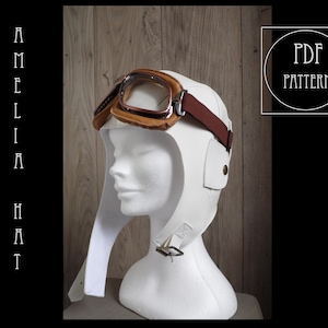Könnte beinhalten: Weißer Lederfliegerhut mit braunem Lederriemen und Brille. Der Hut hat eine Klappe, die heruntergezogen werden kann, um die Ohren zu bedecken. Der Hut befindet sich auf einem weißen Mannequin-Kopf. Der Text "PDF PATTERN" befindet sich in einem Kreis in der oberen rechten Ecke des Bildes.