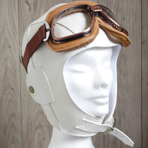 May include: White leather aviator helmet with a brown leather strap and a pair of copper-colored goggles.