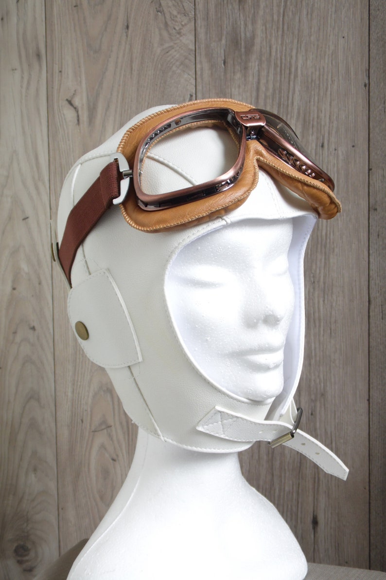 May include: White leather aviator helmet with a brown leather strap and a pair of copper-colored goggles.