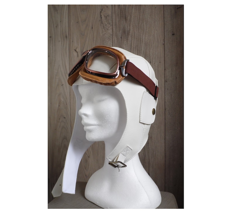 May include: White leather aviator helmet with a brown leather strap and goggles. The helmet has a long flap that can be worn down or up.