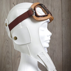 May include: White leather aviator helmet with a brown leather strap and brown leather goggles. The helmet has a flap that covers the neck and chin.