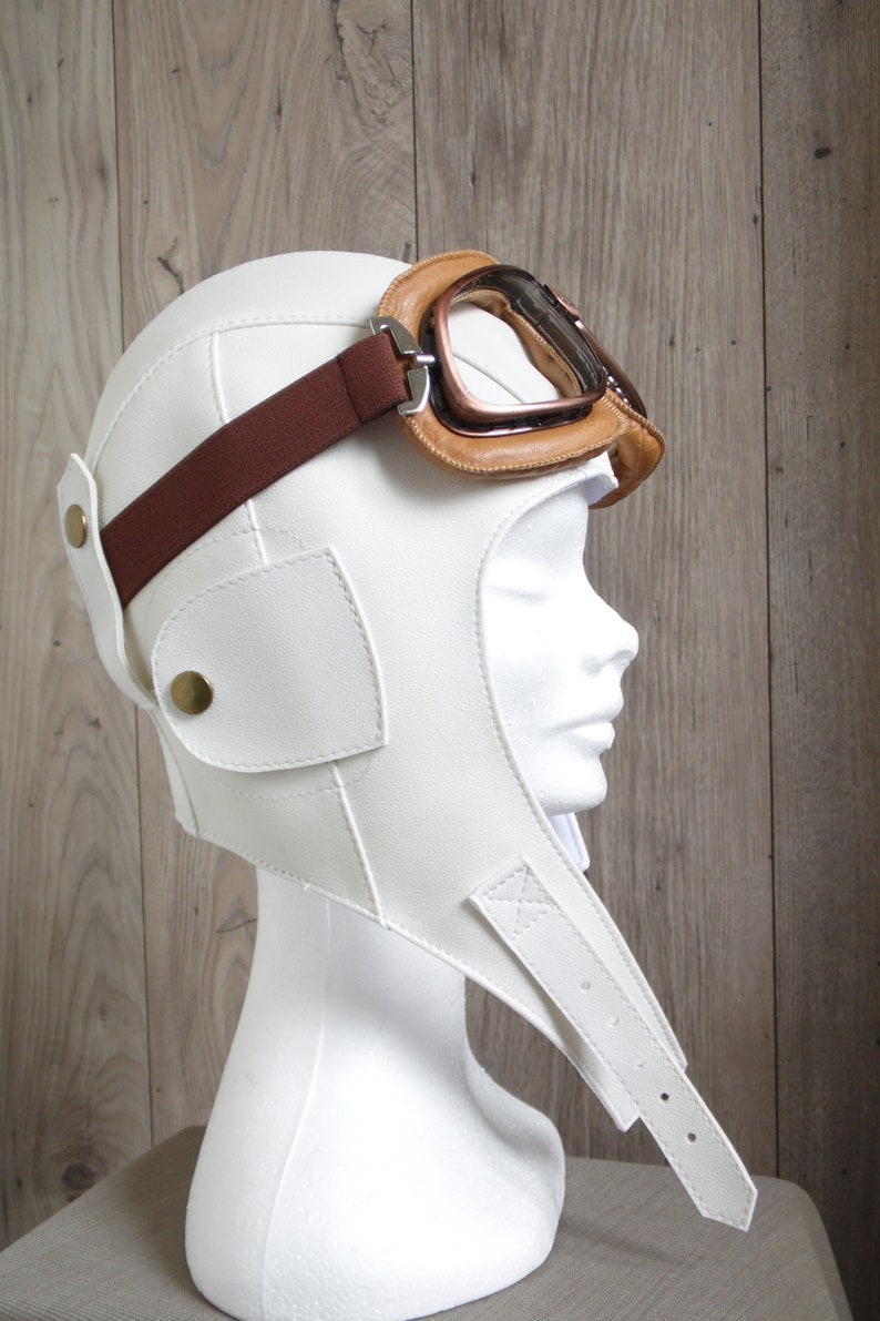 May include: White leather aviator helmet with a brown leather strap and brown leather goggles. The helmet has a flap that covers the neck and chin.