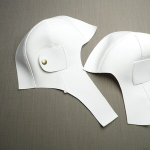 May include: Two white leather helmet pieces with gold buttons. The pieces are laid out on a brown surface.