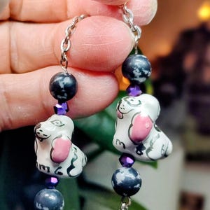 May include: Eyeglass chain featuring white dog-shaped ceramic beads with pink ears and black spots, connected by a silver chain. The chain includes dark gray and purple beads, and a silver paw print charm.