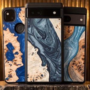 May include: Three smartphone cases featuring a combination of natural light-colored wood burl and swirling blue or dark grey resin. Each case has a black frame and a distinct, organic pattern where the wood grain meets the smooth, marbled resin.