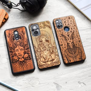 Könnte beinhalten: Drei Handyhüllen aus Holz mit aufwendigen Lasergravuren. Die Hüllen zeigen Bilder einer Gottheit, einer ziegenartigen Figur und einer Anubis-ähnlichen Figur. Die Hüllen sind braun mit schwarzen Rändern, auf einer hellen Oberfläche.